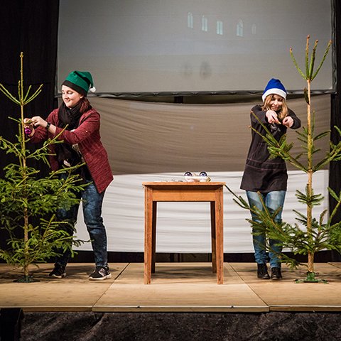 Zwei junge Frauen mit Weihnachtsmützen halten auf einer Bühne Tannenbäume