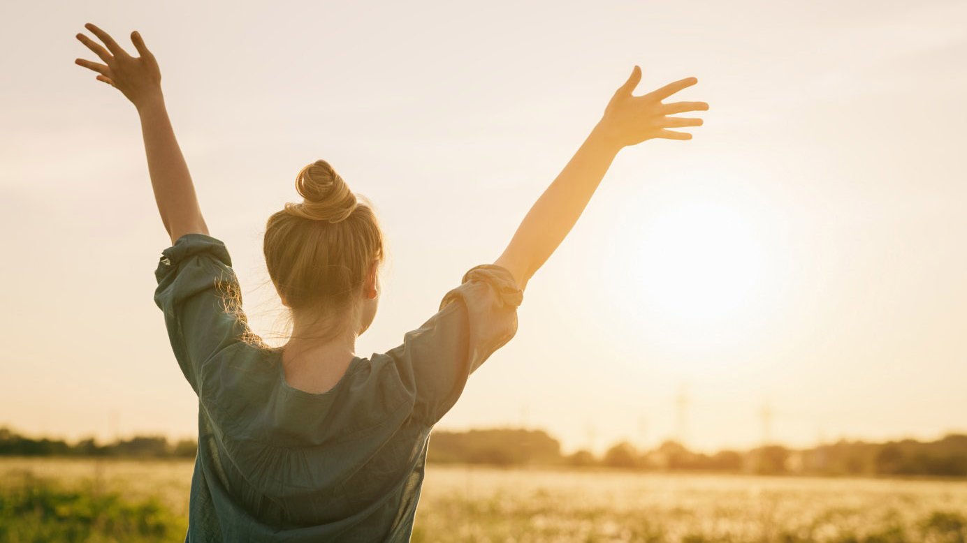 Junge Frau blickt mit erhobenen Armen in die Sonne 