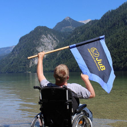 Junger Mann im Rollstuhl steht im Wasser und hält die JuB-Fahne hoch.