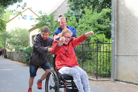 Junge Frau schiebt mit etwas Hilfe von einem Jungen mit Down-Syndrom eine junge Frau im Rollstuhl einen Weg hoch.