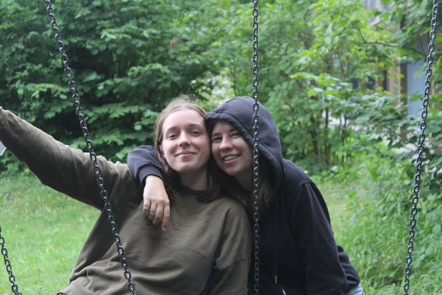 Helene und Anna sitzen auf Schaukeln und lachen Arm in Arm in die Kamera.