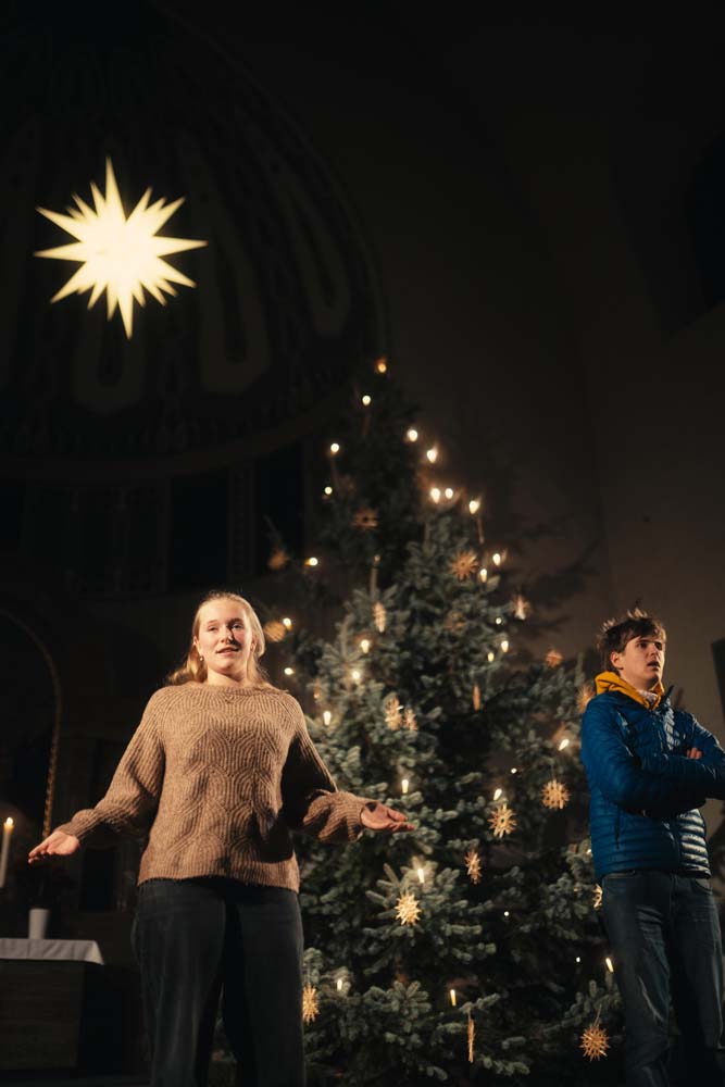 Zwei Personen (Maria und Josef) stehen vor einem geschmückten Weihnachtsbaum. Hinter ihnen leuchtet ein Adventsstern.
