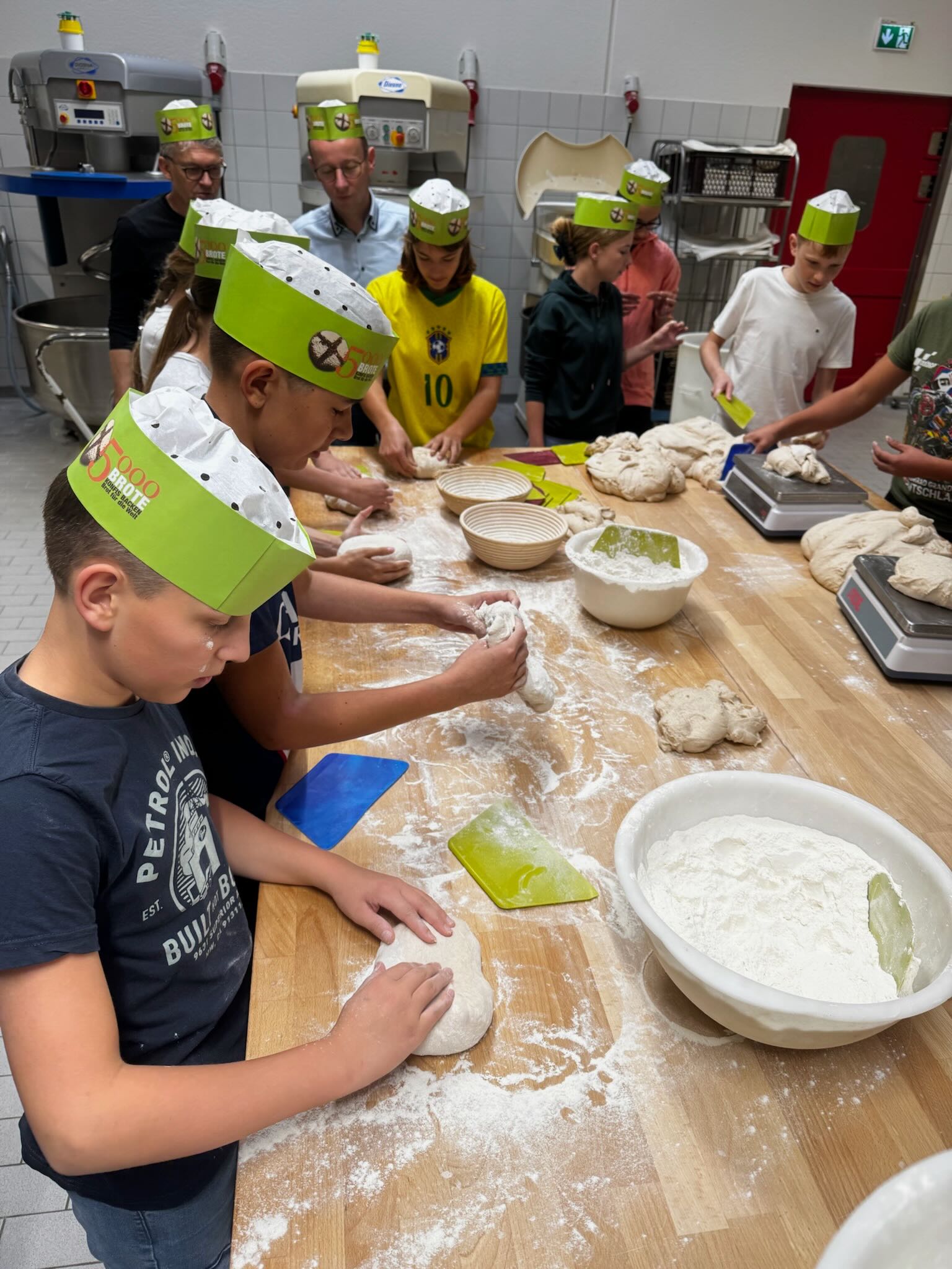 Konfis mit grünen Bäckerschiffchen auf dem Kopf kneten Brot