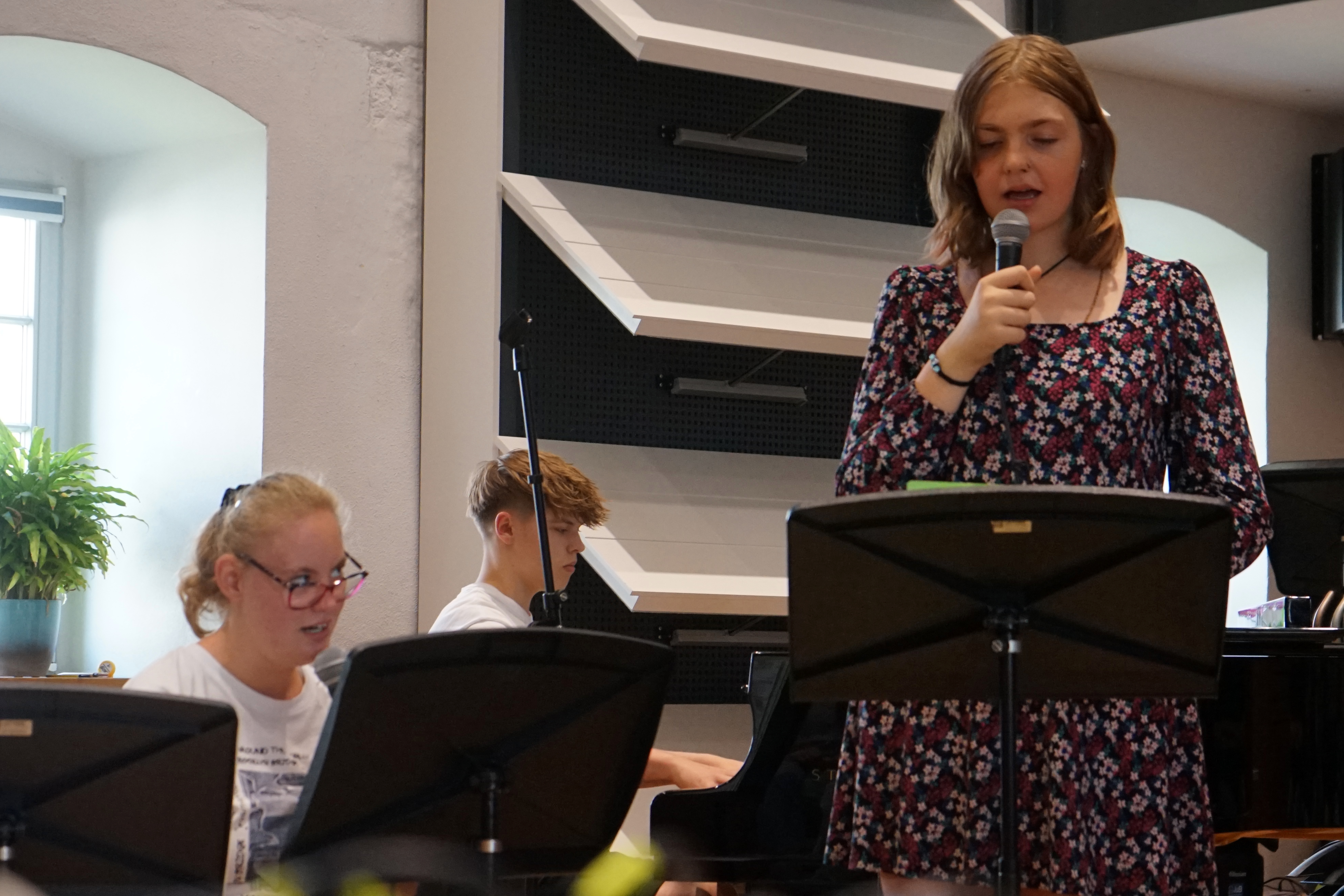 Im Hintergrund sitzt ein Junge, er spielt Klavier. Im Vordergrund sind zwei Frauen zu sehen die Singen.