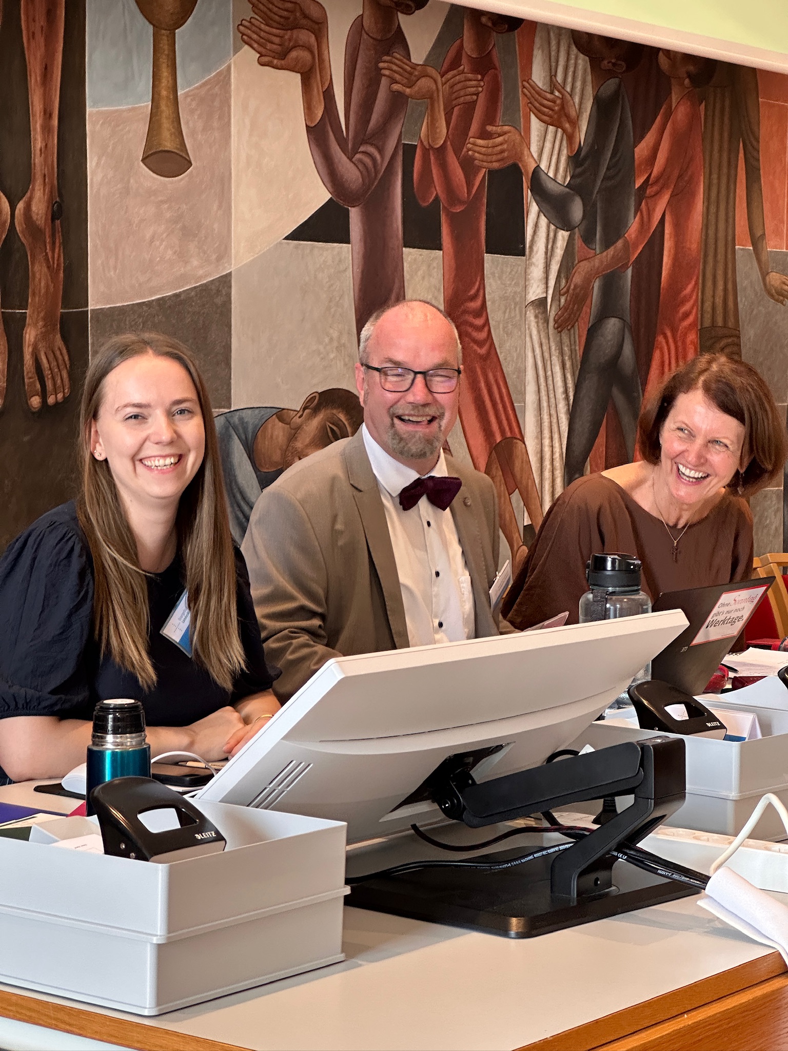 Drei Personen lachen in die Kamera. Sie sitzen an einem Tisch mit Bildschirm und Büromaterial